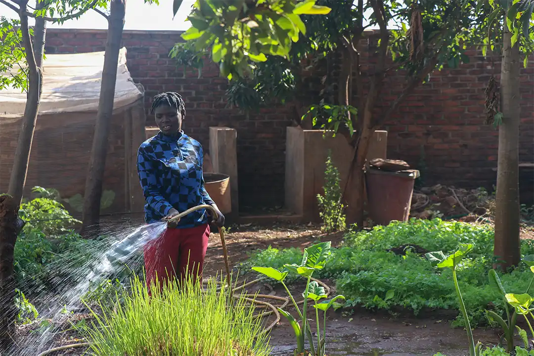 An African lady waters her garden, it has a range of grasses, foliage, and trees